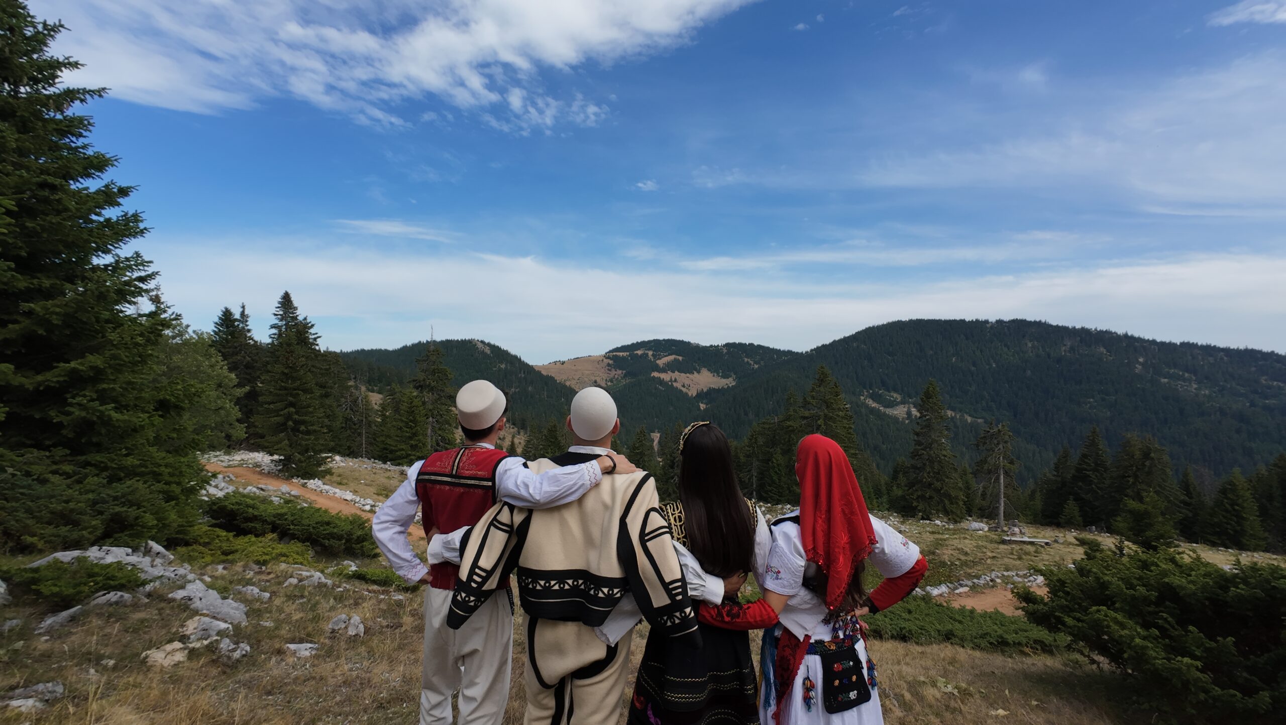 Four Kreshnikët performers in traditional costume, seen from behind looking across a mountain landscape